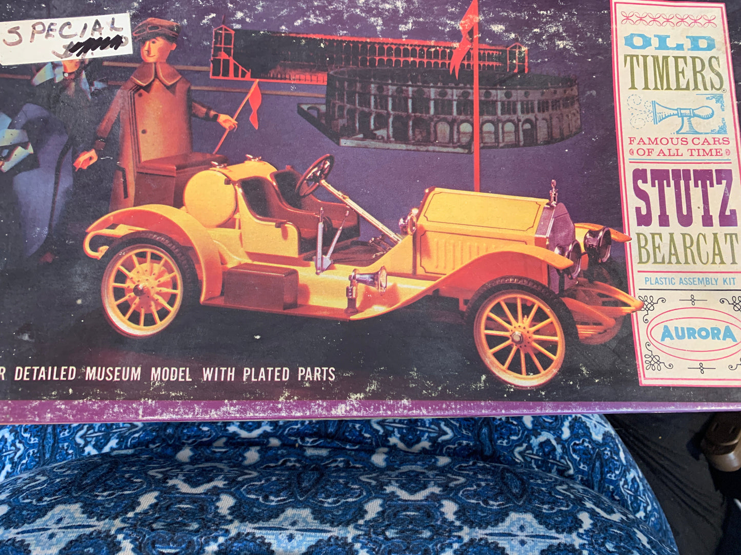 A vintage model car kit box by Stutz, featuring an illustration of a yellow 1910-style open car on a dark illustrated background. The box shows visible wear and tear, with a blue patterned fabric surface visible on the left side.