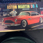 A boxed model kit for an Aston Martin DB 4, photographed on a dark surface next to a keyboard. The box art shows a red classic car and the text 'Aston Martin DB 4' and '1/25 Scale All-Plastic Assembly Kit.