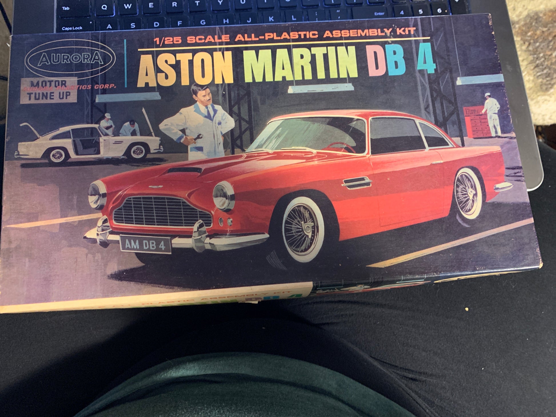 A boxed model kit for an Aston Martin DB 4, photographed on a dark surface next to a keyboard. The box art shows a red classic car and the text 'Aston Martin DB 4' and '1/25 Scale All-Plastic Assembly Kit.