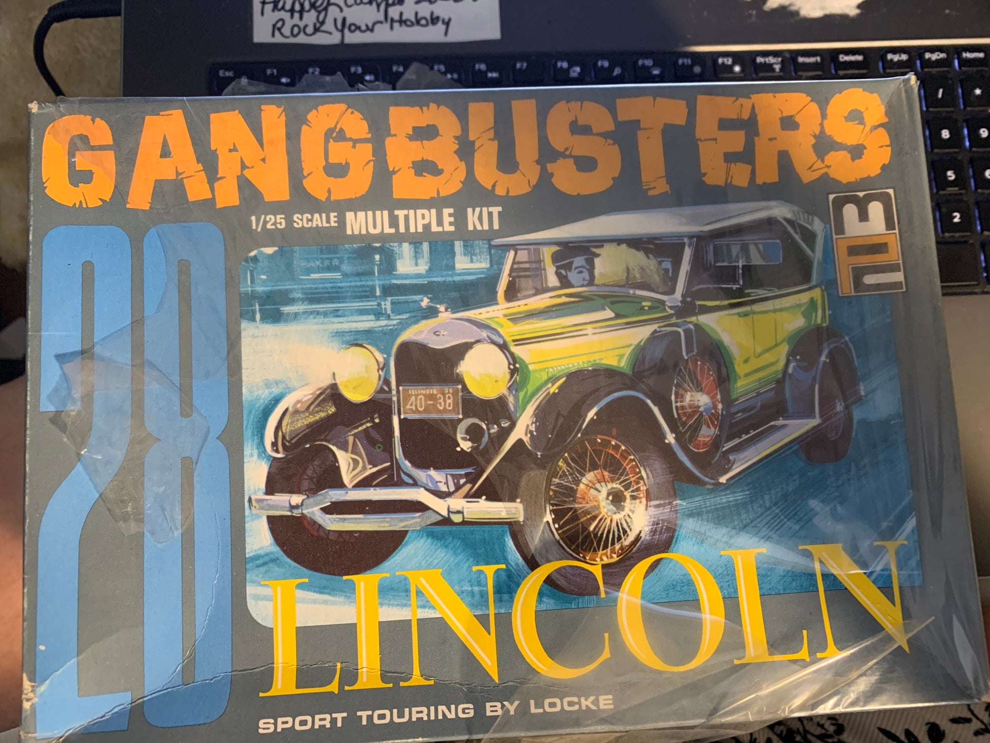 A boxed model kit labeled 'GANGBUSTERS 28 LINCOLN' with a vintage car illustration on the front, photographed on top of a keyboard and laptop.